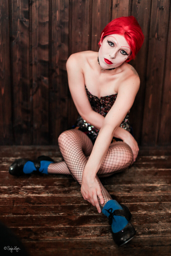 Cosplayer als Columbia aus The Rocky Horror Picture Show sitzt mit überkreuzten Beinen leicht nach vorne gebeugt am Boden.